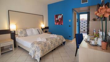 A hotel room with a bed, bedside tables, a desk with a chair, a vase with flowers, and a door leading to a balcony.