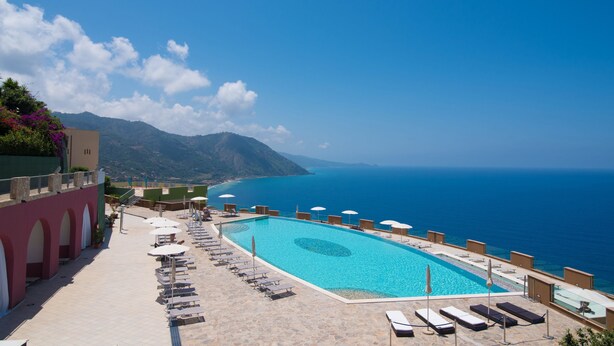 A poolside area with lounge chairs and umbrellas, overlooking a clear blue sea and mountains.