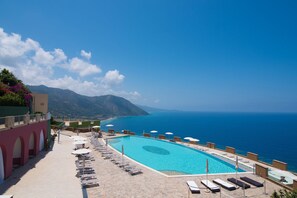 Zona de piscina con sillas de descanso y sombrillas, con vista al mar azul y a las montañas.