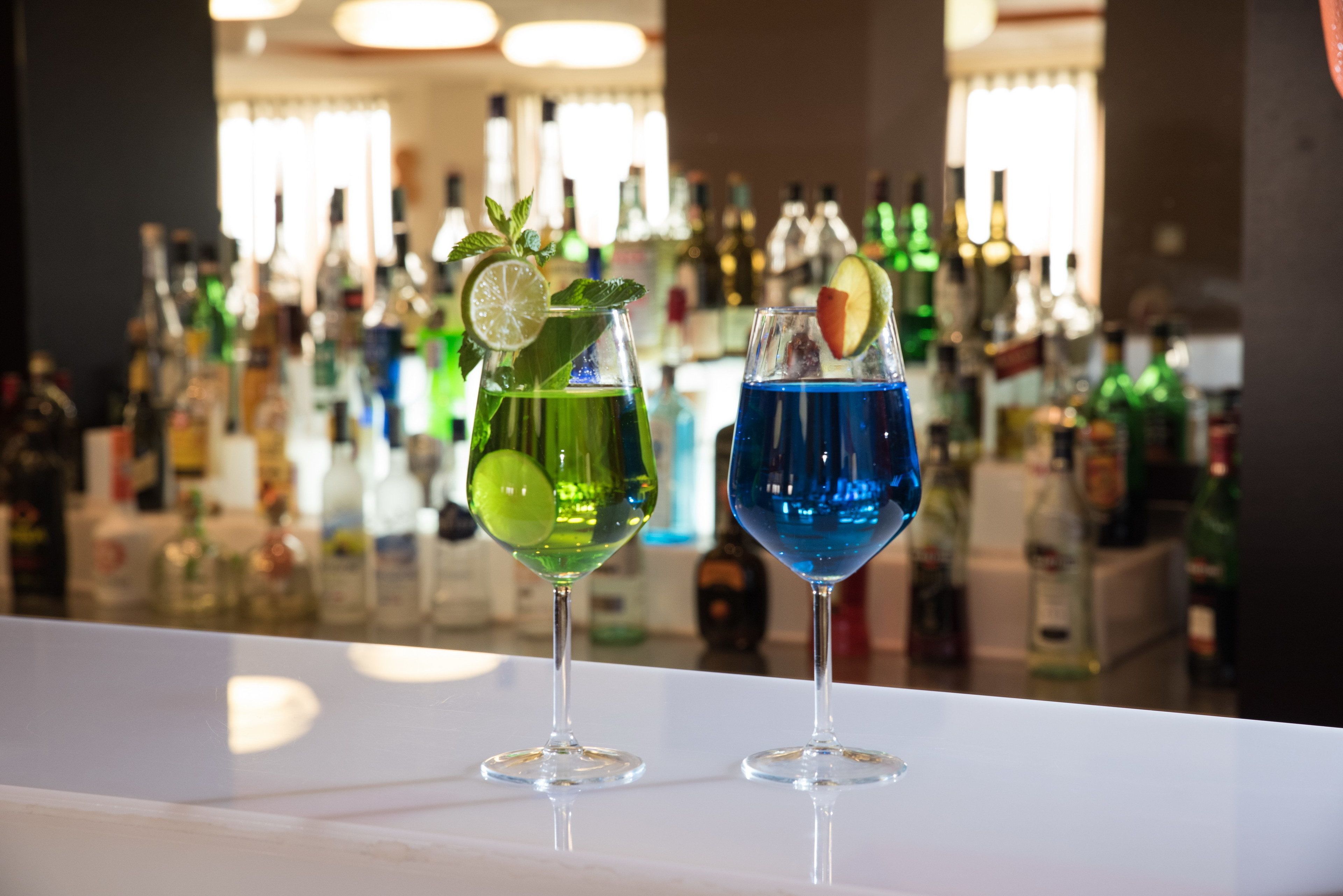 Two cocktails on a bar counter with a background of various bottles.