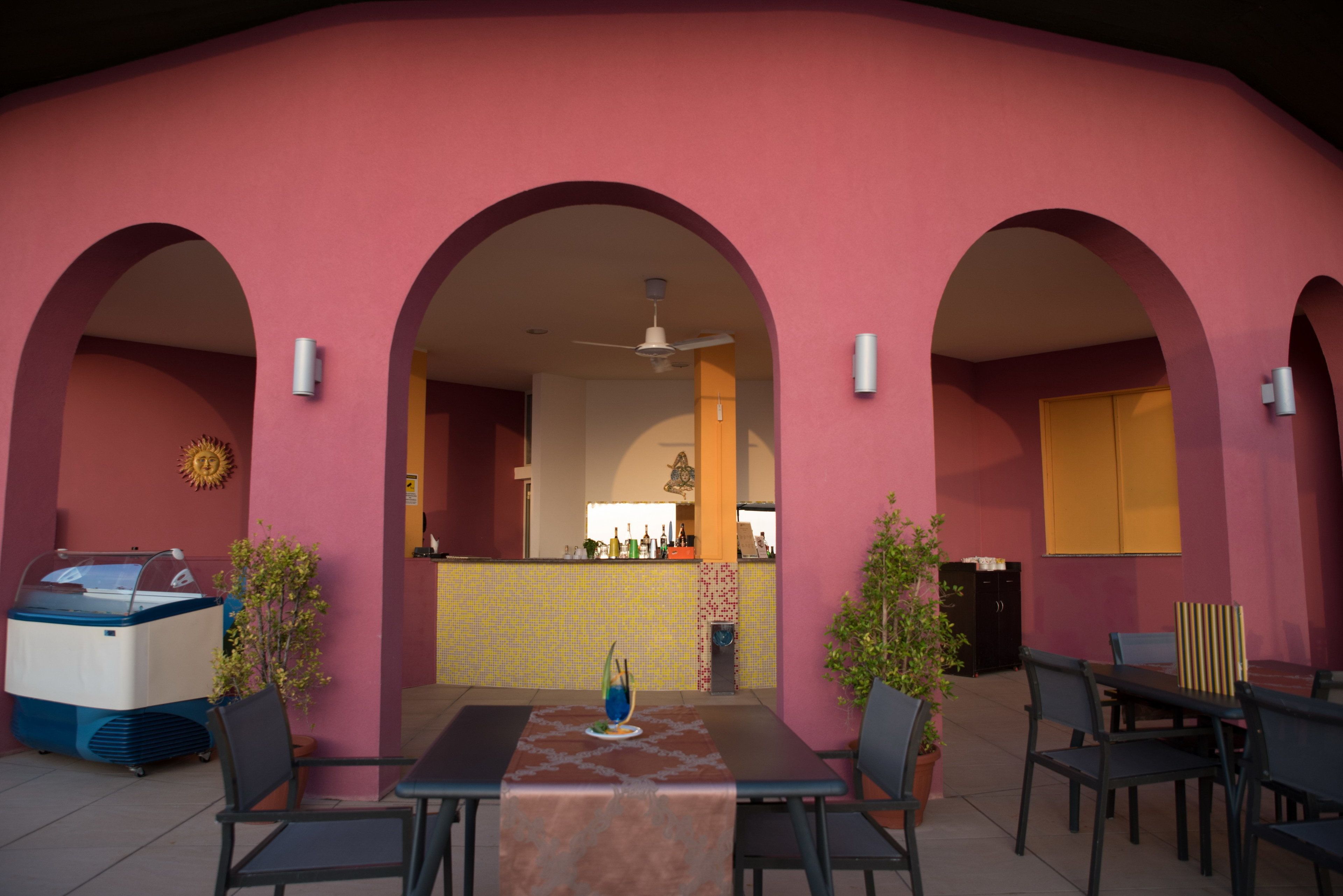 Un restaurante con fachada rosa, mesas al aire libre y una zona de bar central.