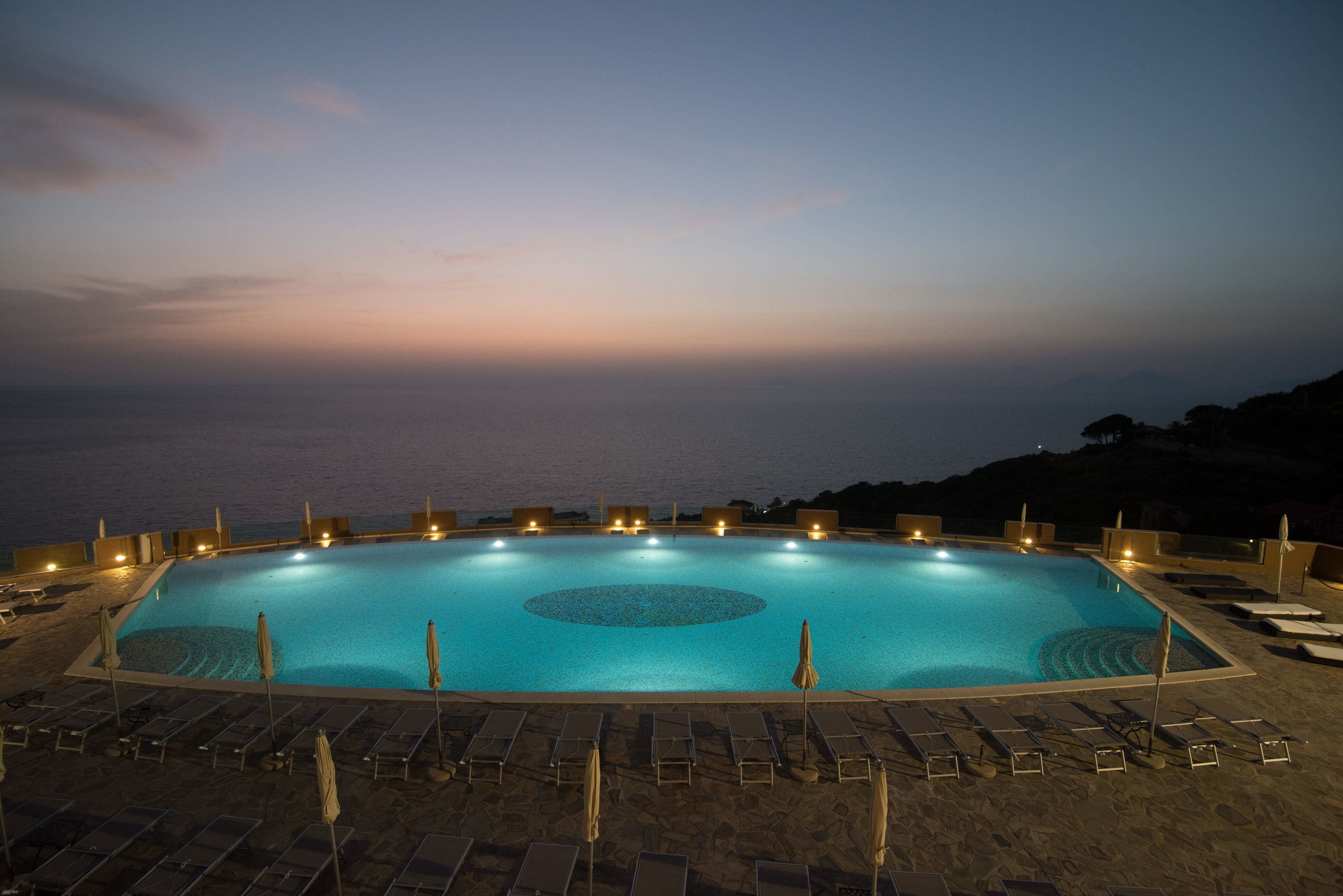 Piscina rectangular con un centro circular, rodeada de sillas de descanso y sombrillas, con vistas a una masa de agua.