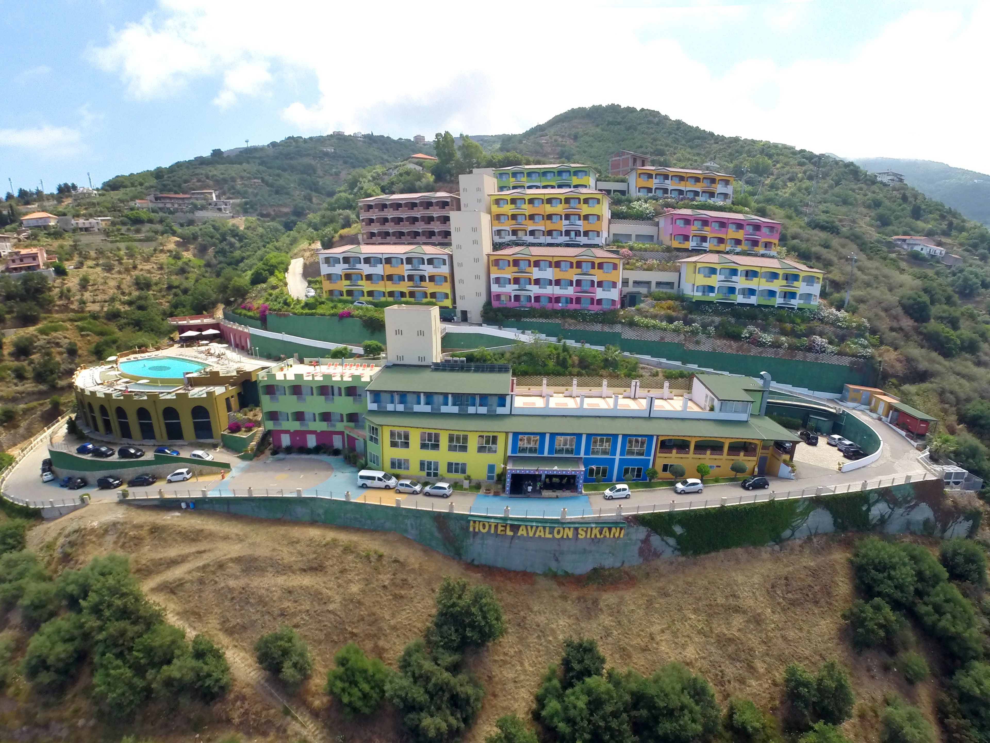 Un hotel de varios pisos con piscina, situado en una colina rodeada de vegetación.
