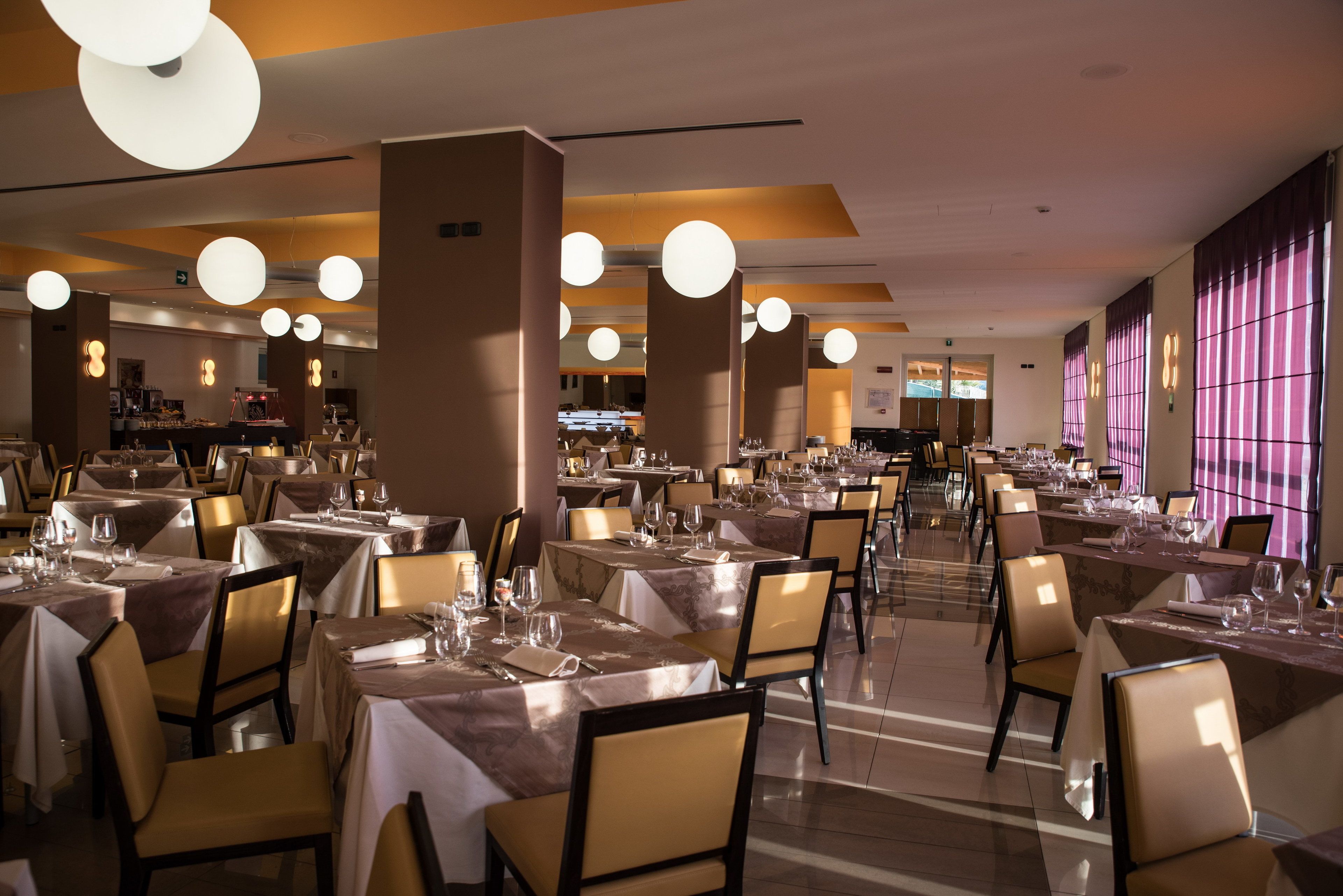 A restaurant with multiple tables set for dining, featuring a modern interior with pendant lighting and a view of the kitchen area.