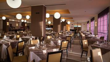 A restaurant with multiple tables set for dining, featuring a modern interior with pendant lighting and a view of the kitchen area.