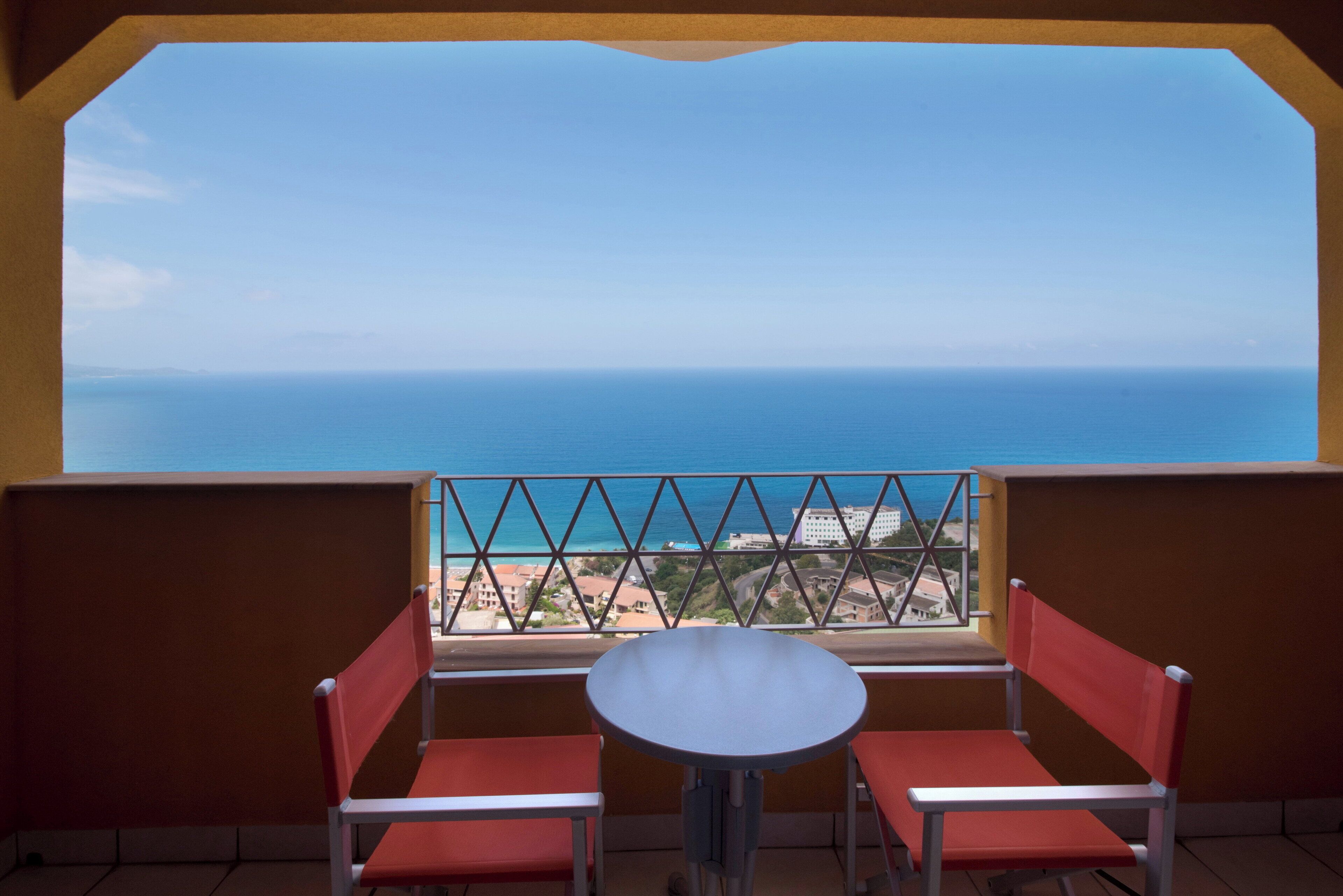 Un balcón con dos sillas rojas y una pequeña mesa redonda, con vistas a la costa, edificios y un cielo despejado.