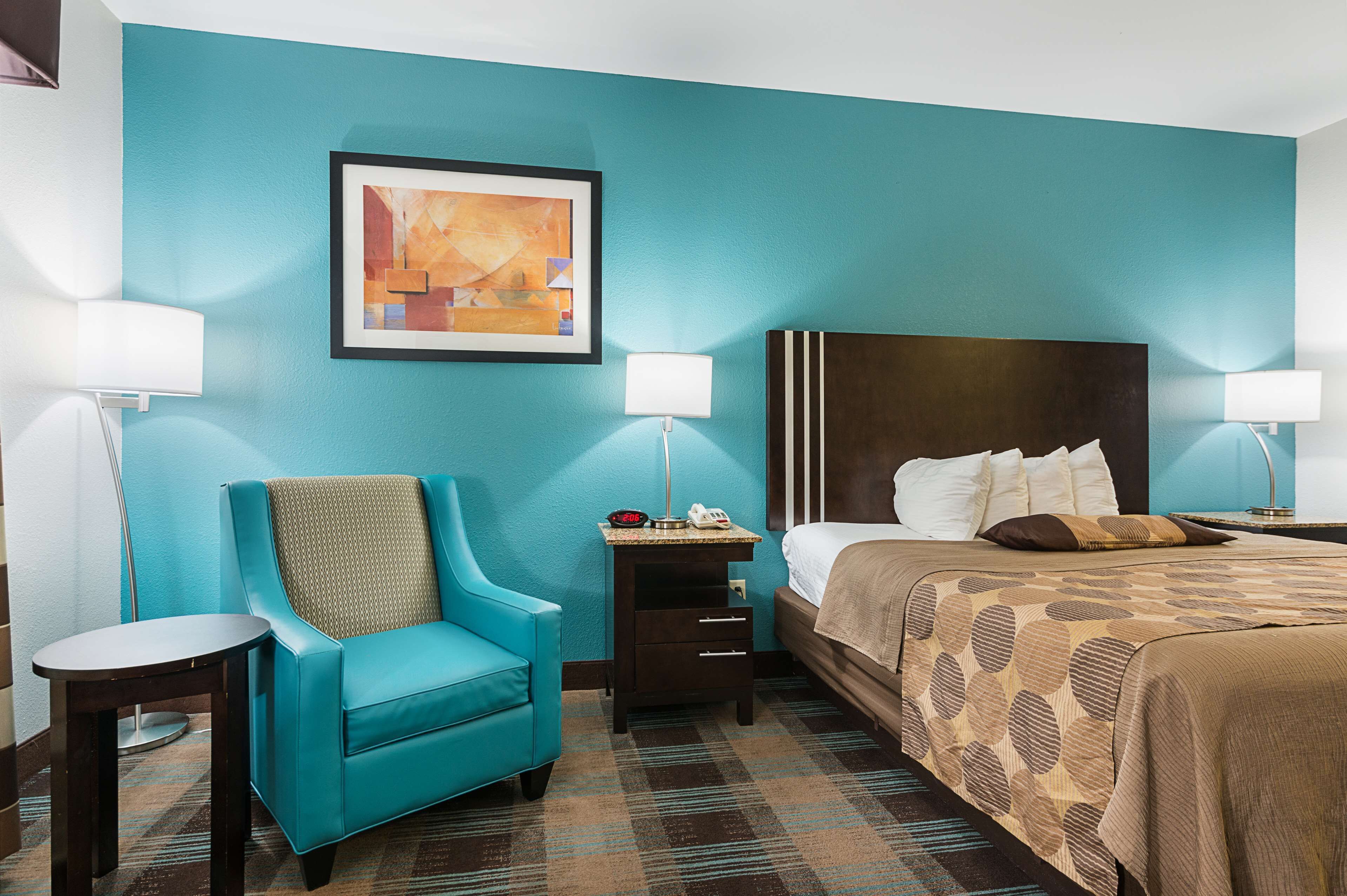 A hotel room with a teal wall, a bed with a patterned bedspread, a blue armchair, a small table, and a nightstand with a lamp.