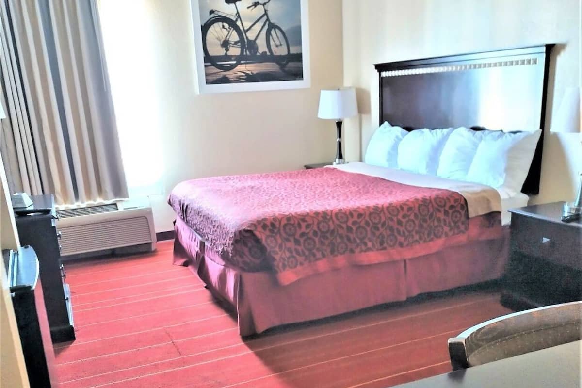 A hotel room with a bed, a desk, a chair, a lamp, a painting, and a window with curtains.