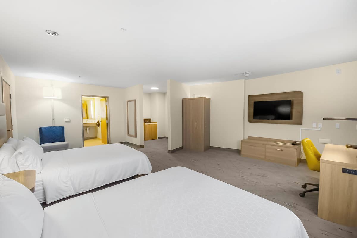 Quarto de hotel com duas camas, uma escrivaninha, uma cadeira, guarda-roupa, televisão e banheiro visível por uma porta aberta.