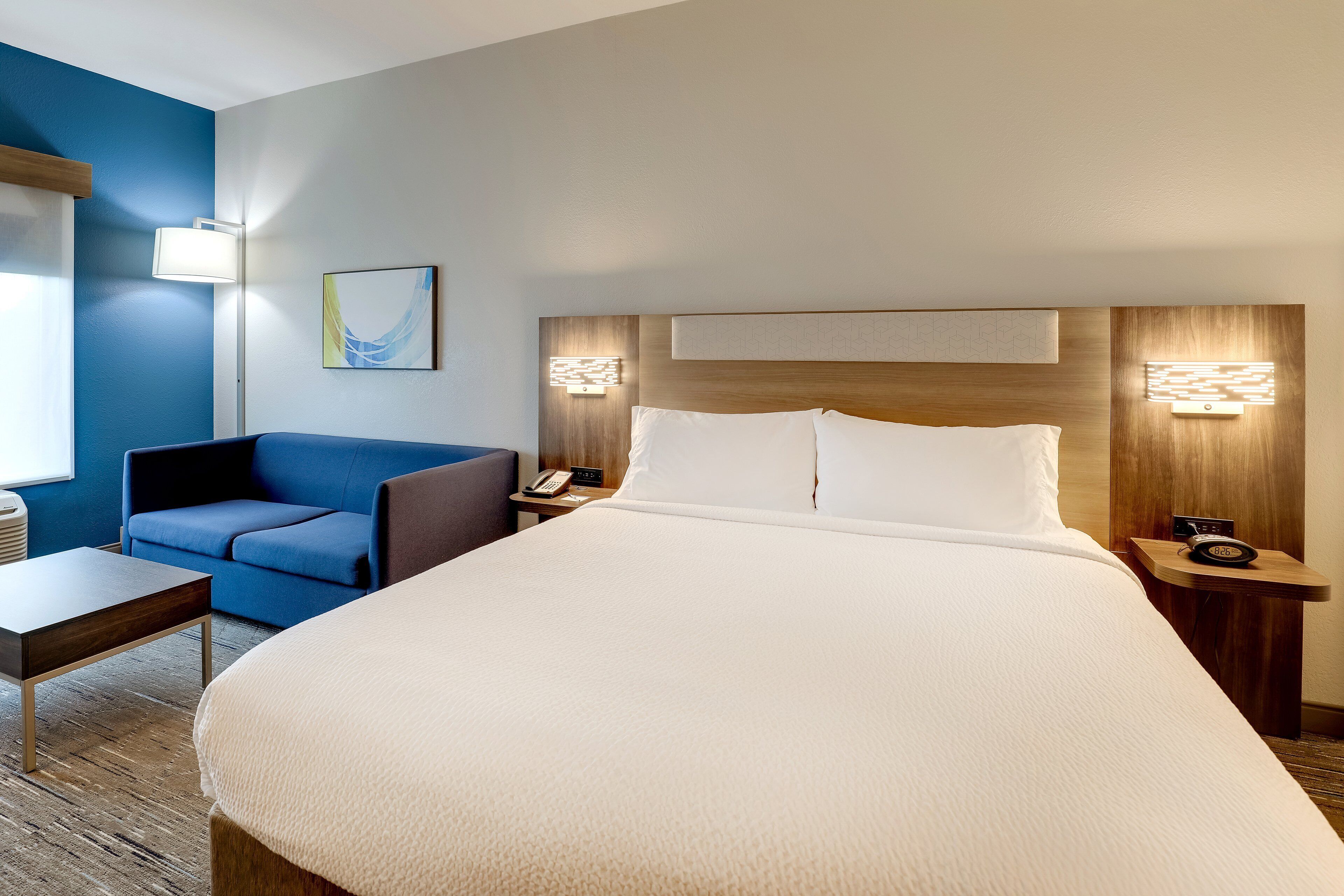 A hotel room with a large bed, a blue armchair, a bedside table with a lamp, and a wall-mounted telephone.