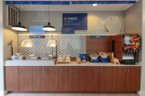A breakfast station with a sign reading "EXPRESS START BREAKFAST" and a sign for "EGG-XACTLY". There are bowls of fruit and a coffee machine.