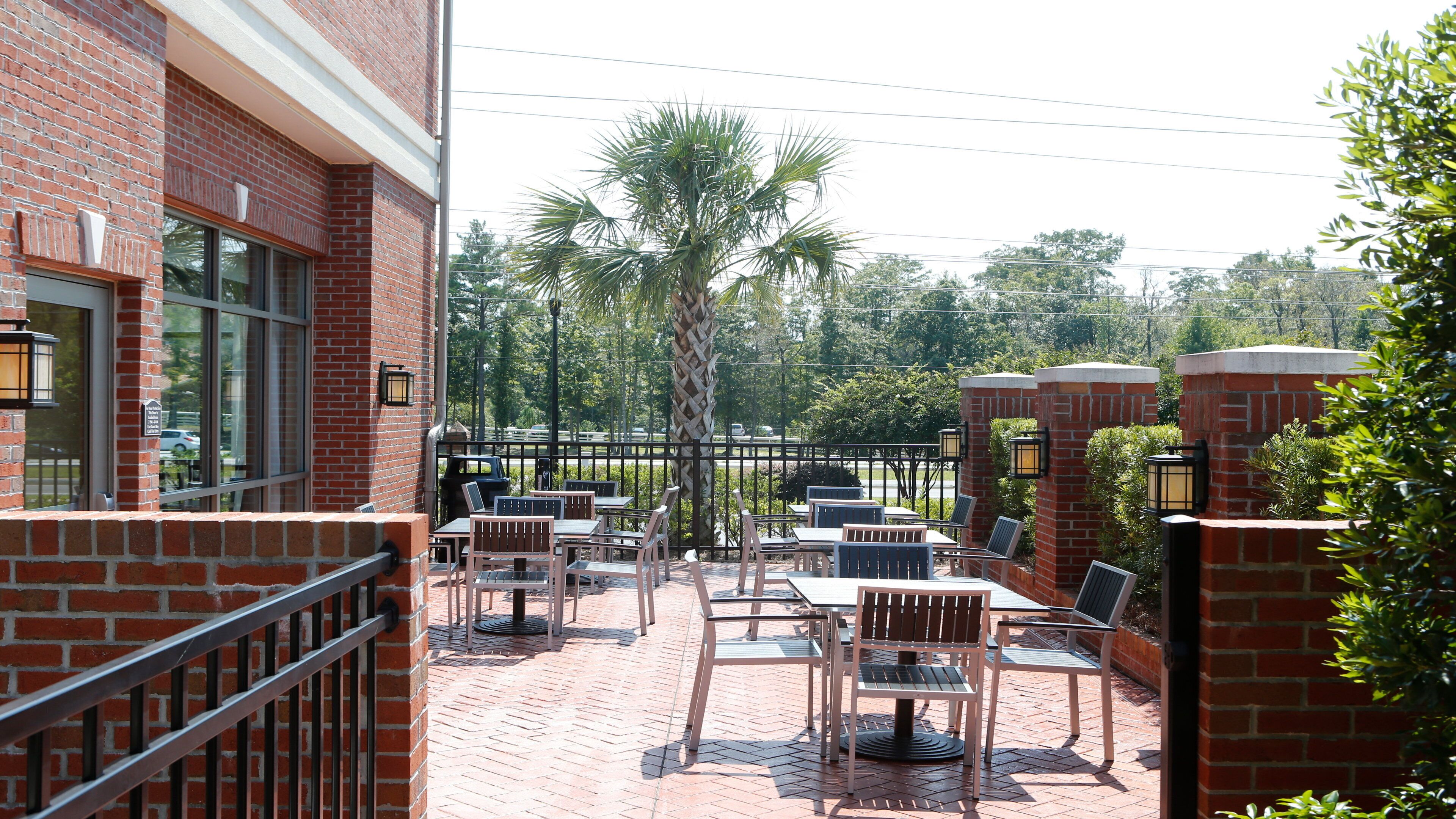 An outdoor patio with brick walls, tables, and chairs, surrounded by a fence and palm trees.