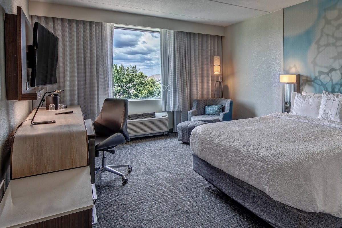 Ein Hotelzimmer mit einem Bett, einem Schreibtisch mit Computer, einem Sessel, einem Fernseher und einem Fenster mit Vorhängen.