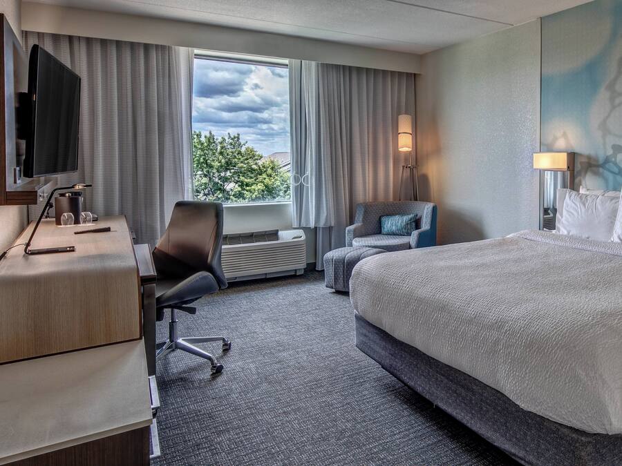 Habitación de hotel con cama, escritorio con computadora, silla, televisor y ventana con cortinas.
