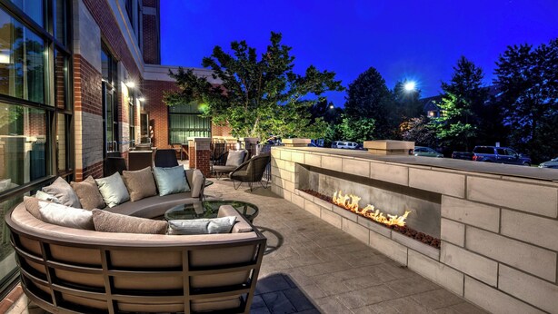 An outdoor seating area with a curved sofa and chairs, a fire feature, and a view of the surrounding trees and parked cars. - Courtyard by Marriott Dulles Airport Herndon (Herndon)