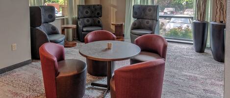 A modern lounge area with a round table, several upholstered chairs, and large windows offering a view of the street.