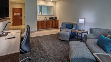 A modern hotel room with a sofa, armchair, desk, and a small kitchenette.