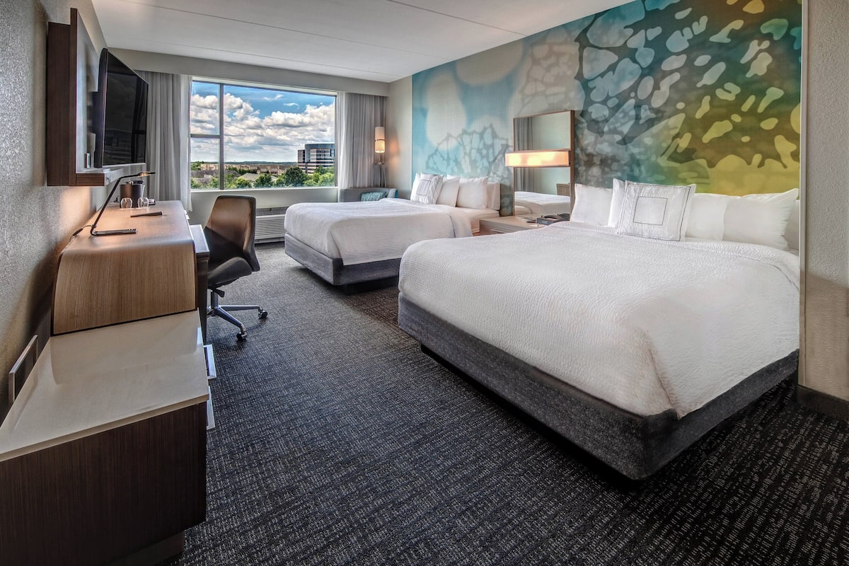 Ein Hotelzimmer mit zwei Betten, einem Schreibtisch, einem Sessel, einem Fernseher und einem großen Fenster mit Blick auf die Stadt.