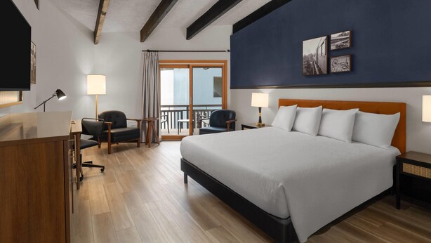 Habitación de hotel con una cama grande, un escritorio con silla, un balcón con vistas y un televisor montado en la pared.