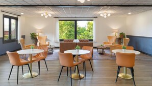 Una sala de conferencias moderna con mesas redondas y sillas anaranjadas, ventanas amplias con vistas a zonas verdes, y un techo con diseño de cuadrícula y luces colgantes.