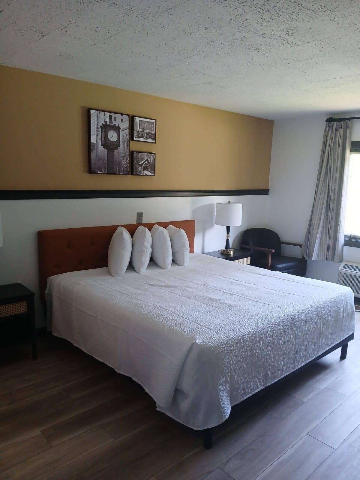 A hotel room with a large bed, a wooden headboard, a nightstand, a desk, and a chair. There are framed pictures on the wall above the bed.