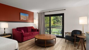 Habitación de hotel con un sofá rojo, una mesa redonda de madera, un sillón negro, una cama y un ventanal con vistas a zonas verdes.