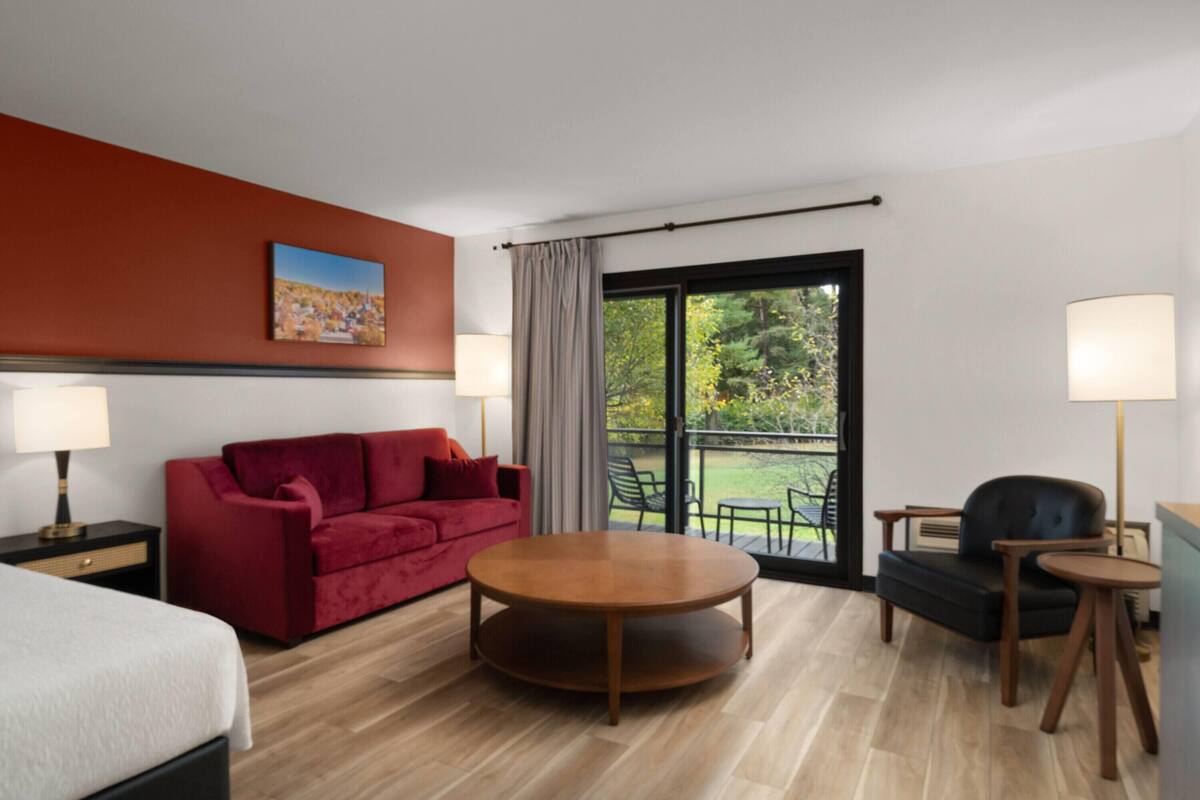 A hotel room with a red sofa, a round wooden table, a black armchair, a bed, and a large window with a view of greenery.