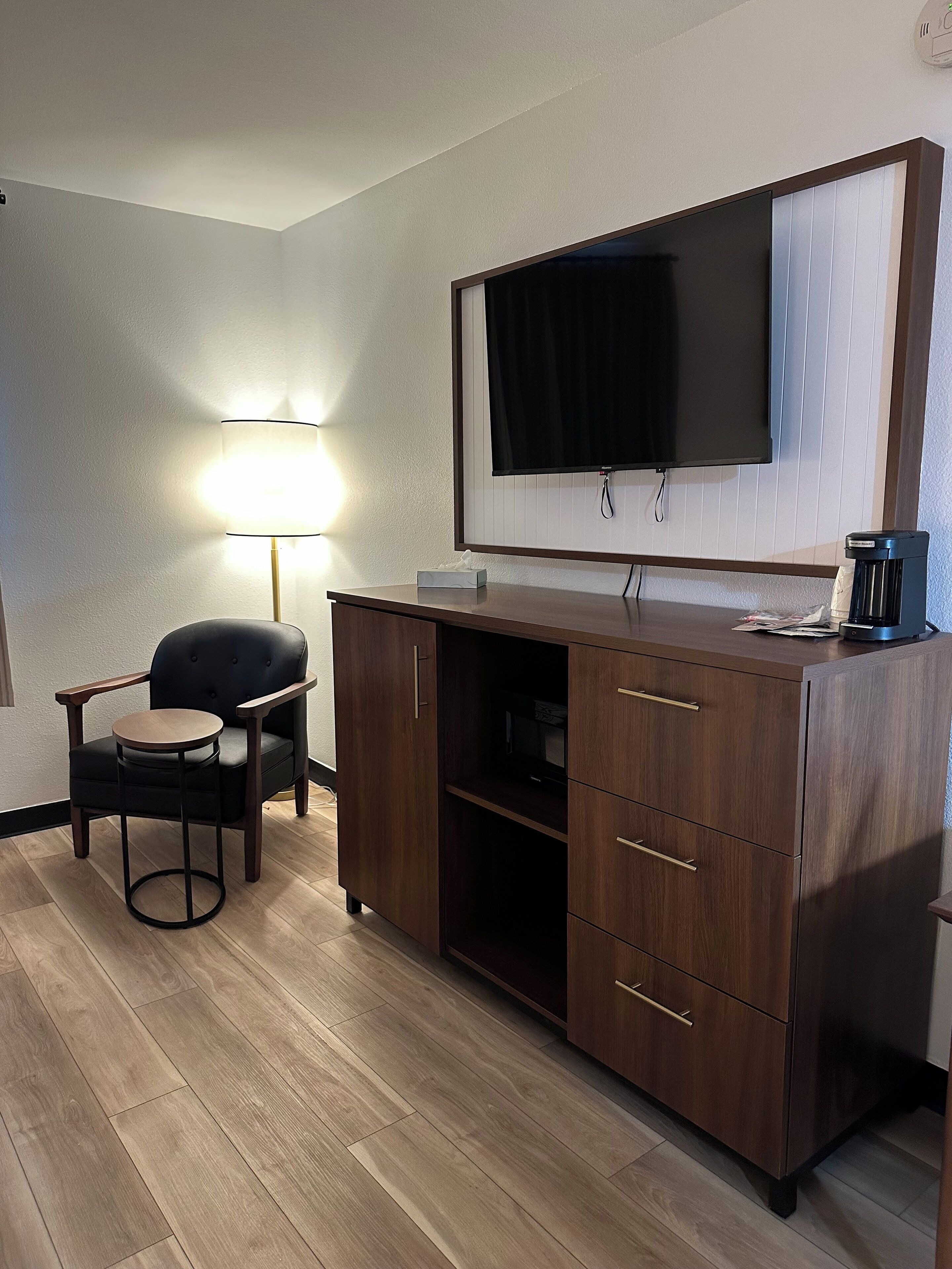 Una habitación de hotel con un escritorio de madera, una silla, un televisor montado en la pared y una mesita de noche con lámpara.