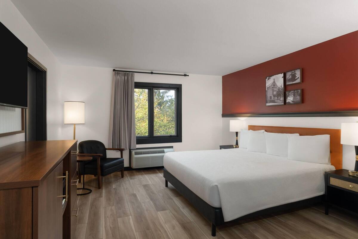 A hotel room with a large bed, a desk, a chair, a window with curtains, and a wall with framed pictures.