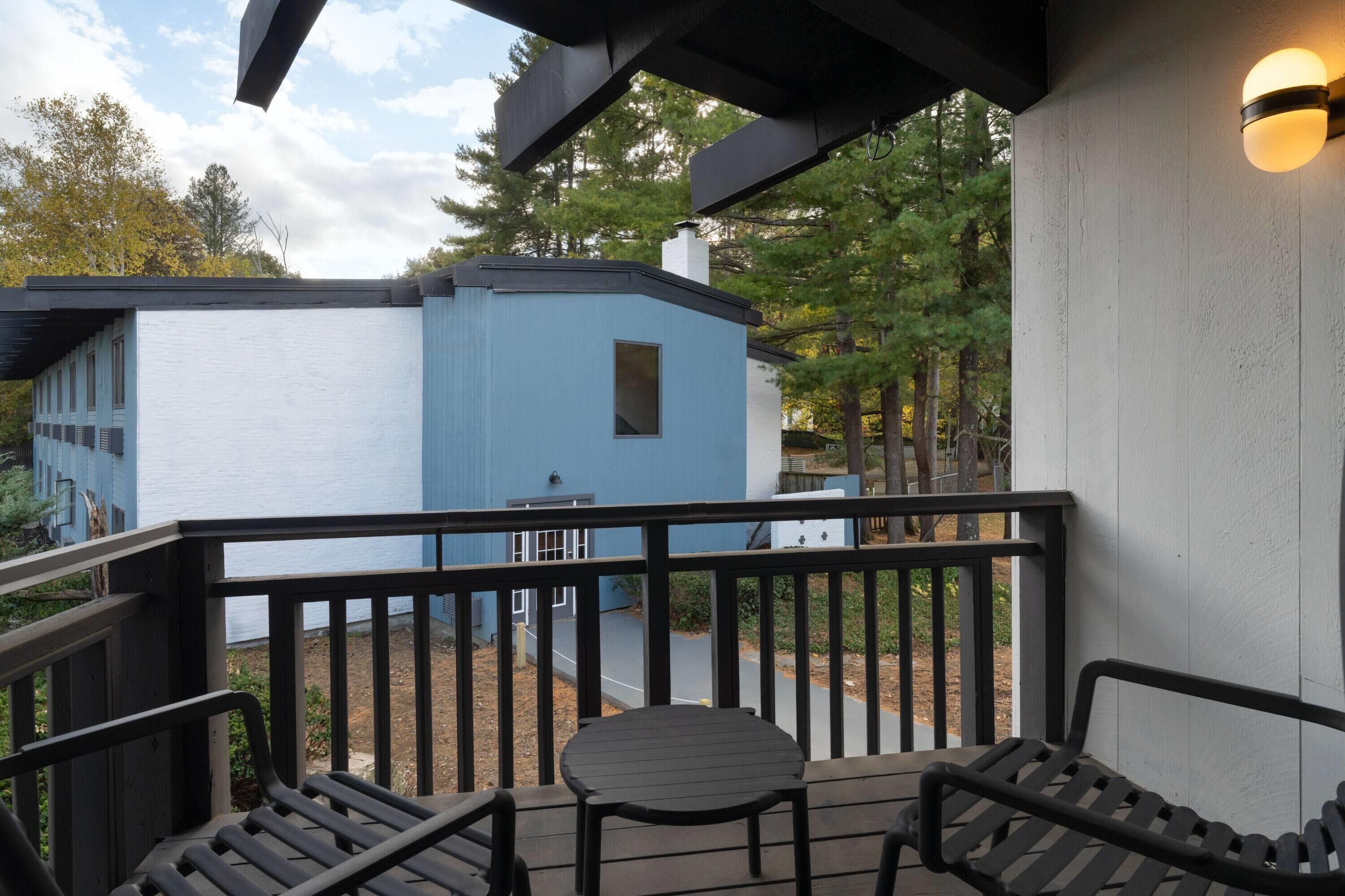 A balcony with a table and two chairs, overlooking a blue building and a forested area.