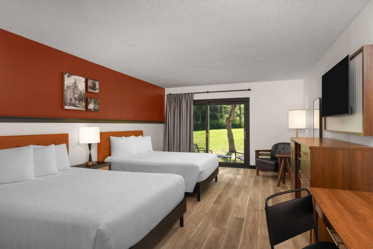 A hotel room with two beds, a desk, a chair, a TV, and a window with a view of greenery.