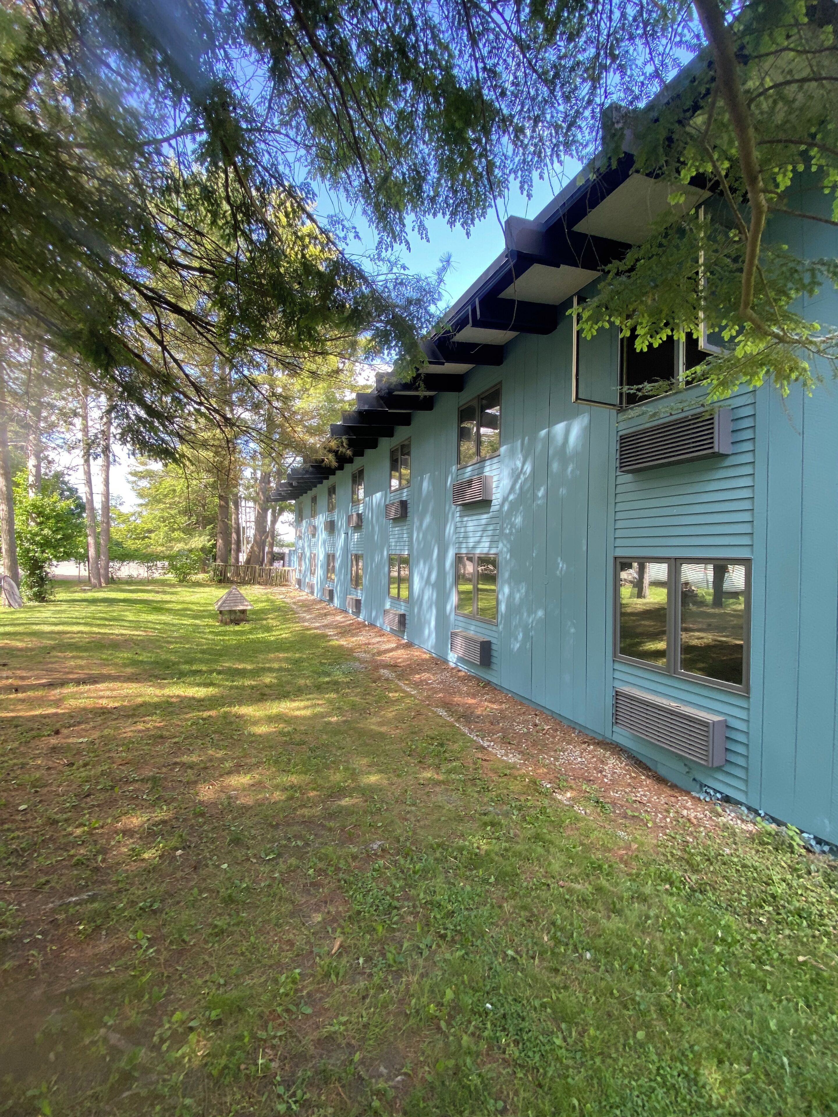 A row of light blue buildings with multiple windows and air conditioning units, surrounded by a grassy area and trees.