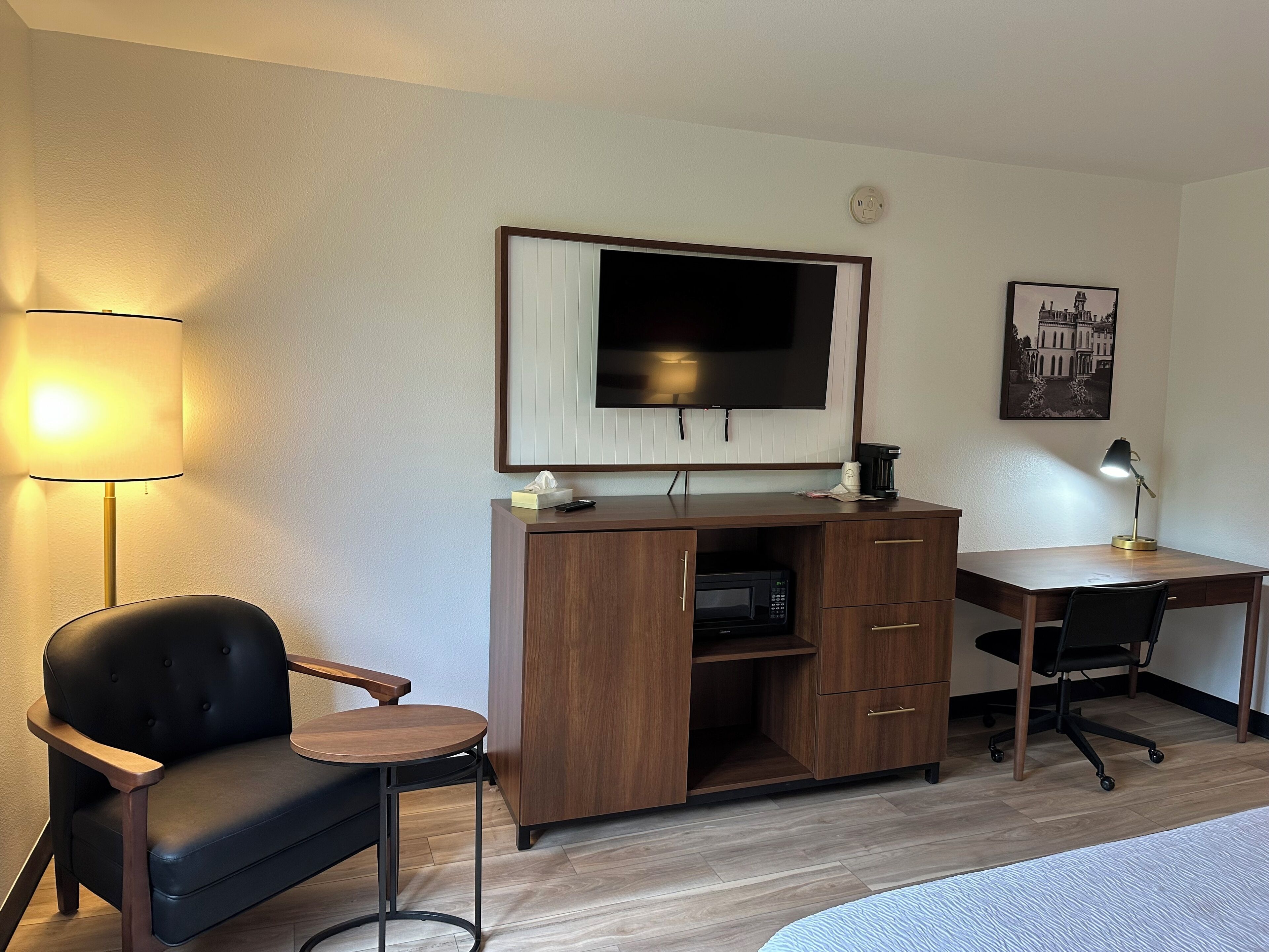 A hotel room with a wooden desk, a black leather chair, a television, a coffee maker, and a lamp.