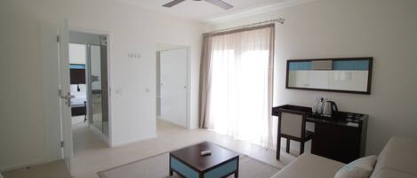 A living room with a sofa, a coffee table, a ceiling fan, a desk with a chair, and a large window with curtains.