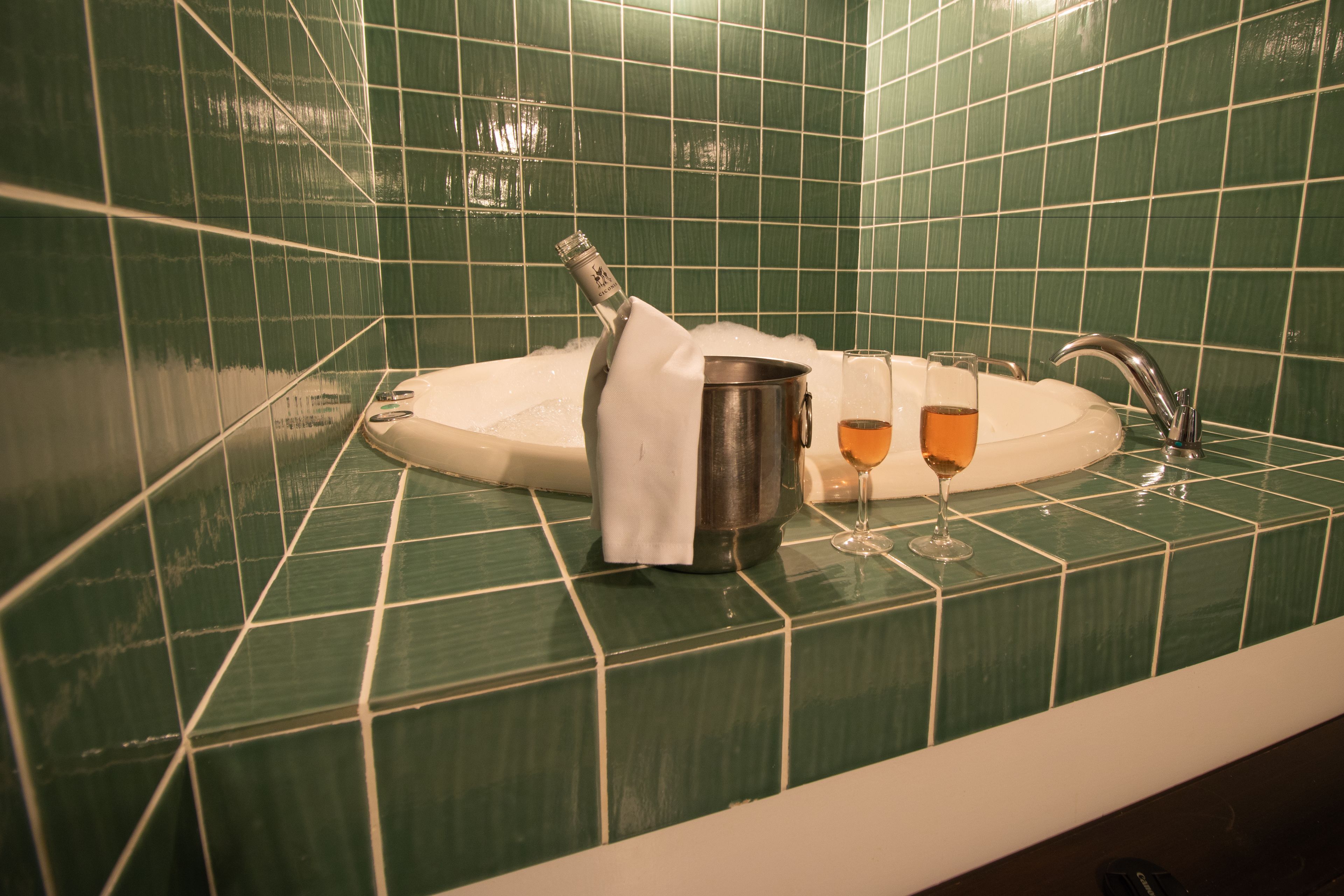 Une salle de bain aux carreaux verts, une baignoire, une bouteille de champagne et deux verres remplis d’un liquide doré.