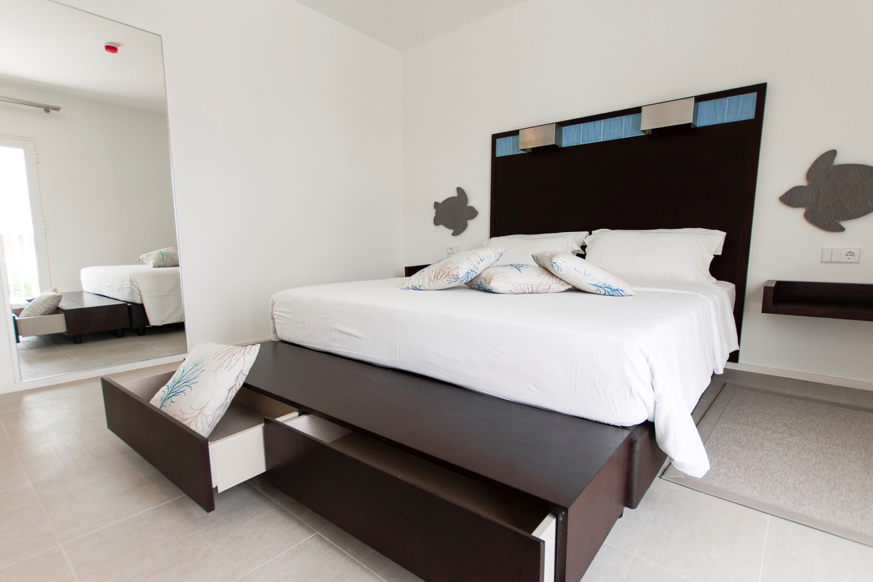A modern bedroom with a large bed, a bench at the foot of the bed, a small desk, and a turtle wall decoration.
