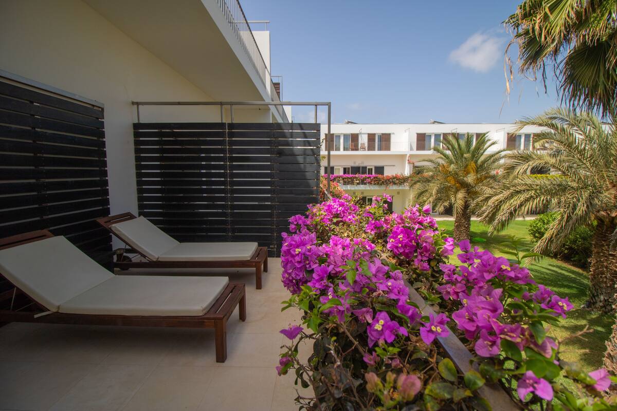 A balcony with two wooden lounge chairs and a black sunshade, overlooking a garden with purple flowers and palm trees.