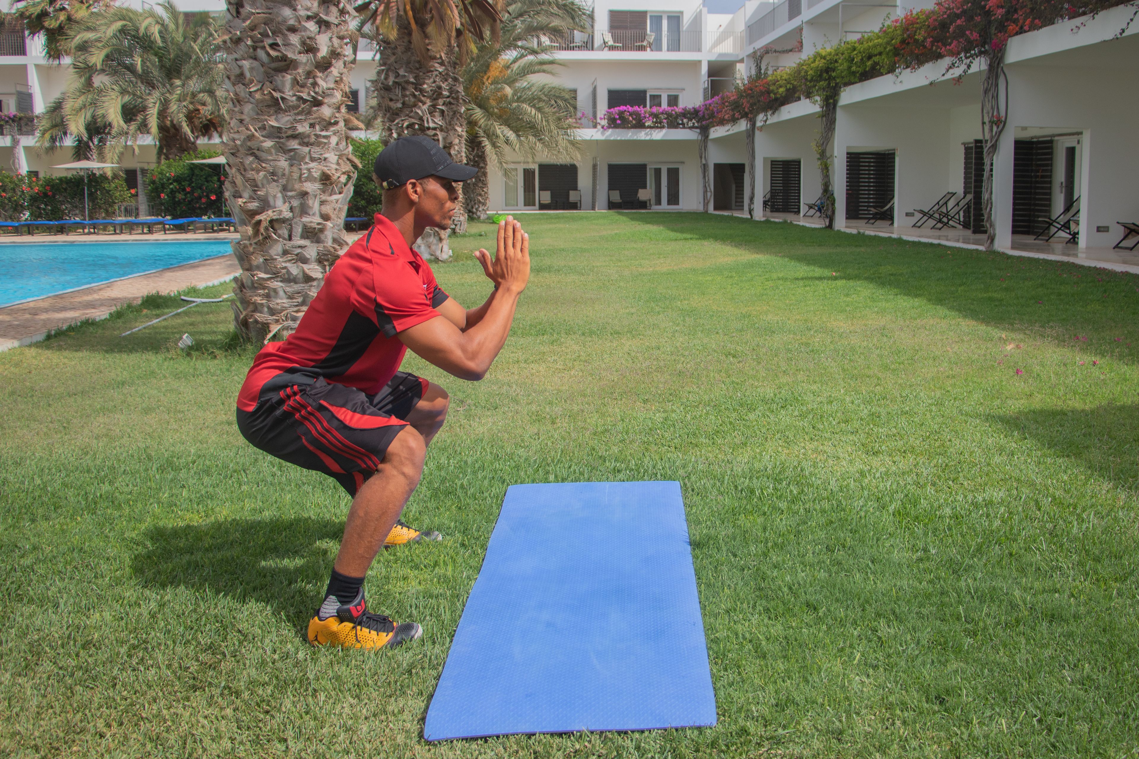 A man performing squats on a blue mat in a courtyard with a pool and palm trees.