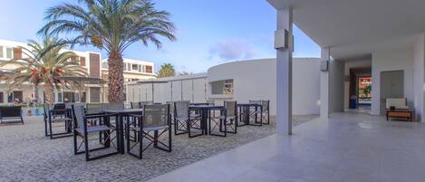 An outdoor patio area with tables and chairs, palm trees, and a modern building in the background.