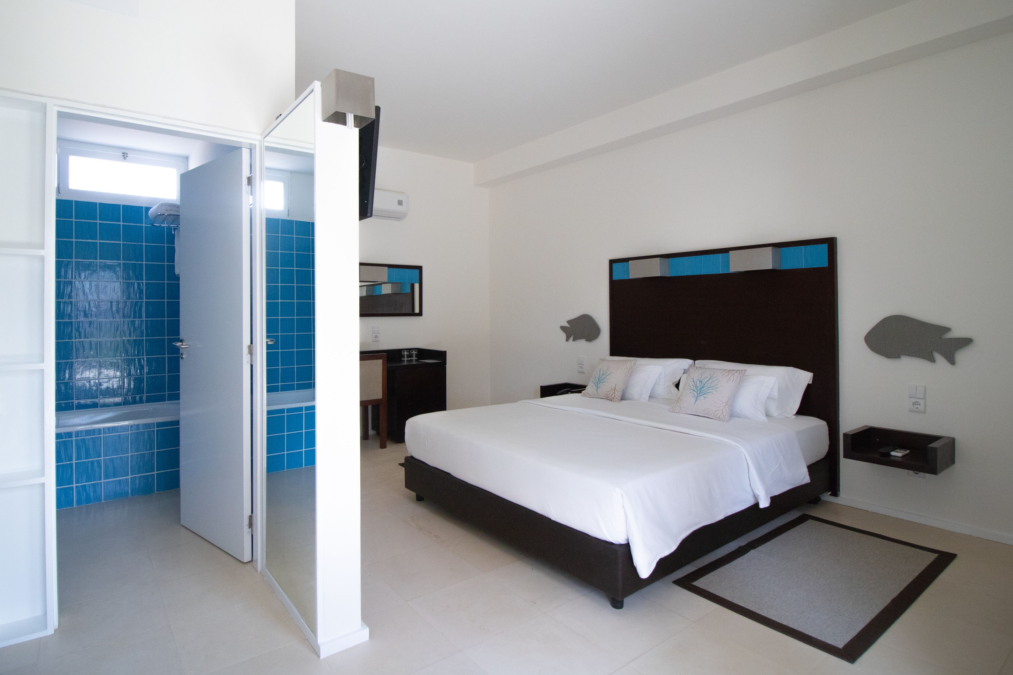 A bedroom with a large bed, a bedside table, a nightstand, a wall-mounted shelf, and a bathroom with blue tiles and a shower.