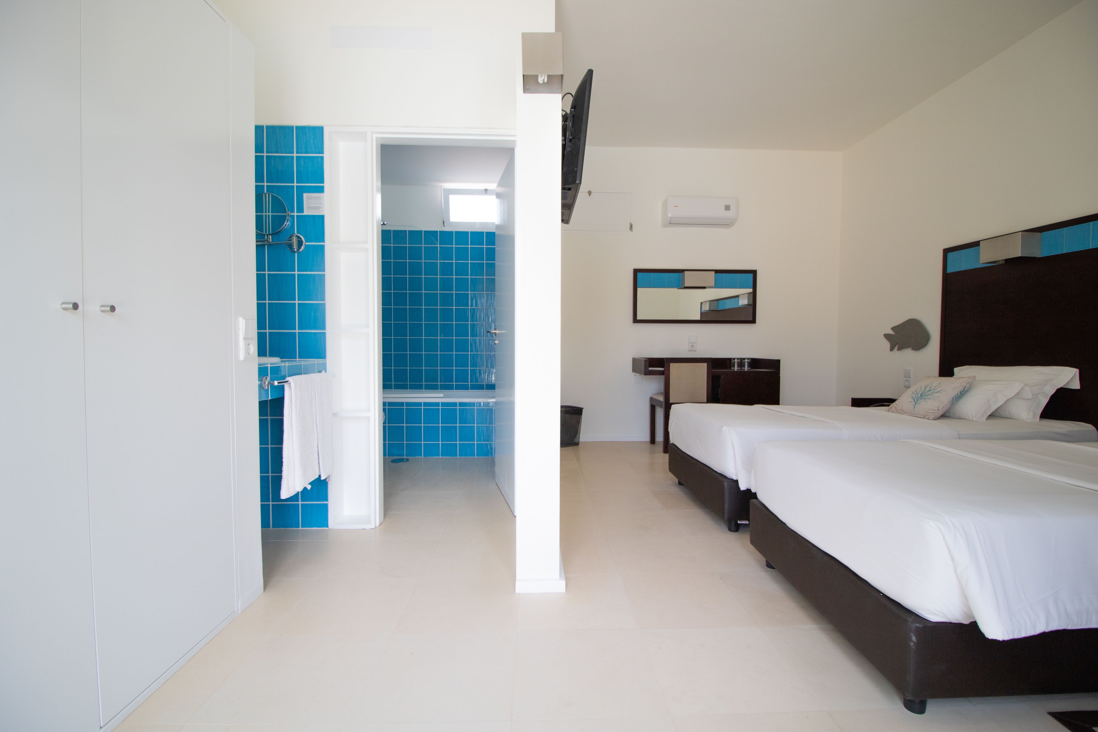 A hotel room with two beds, a bathroom with blue tiles, a flat-screen TV, and a small table.