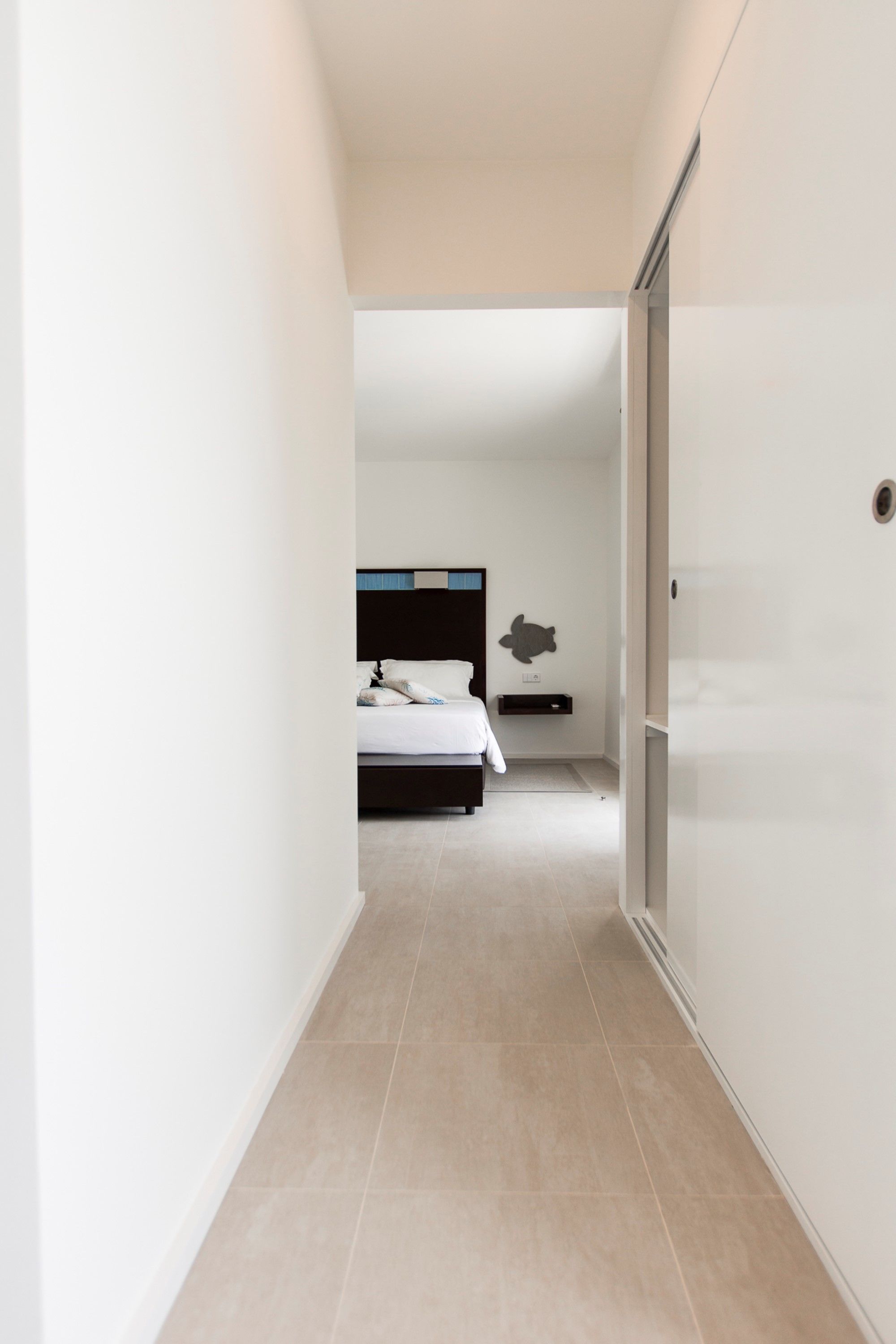 Un couloir étroit mène à une chambre qui contient un lit, une table de chevet et une étagère fixée au mur.