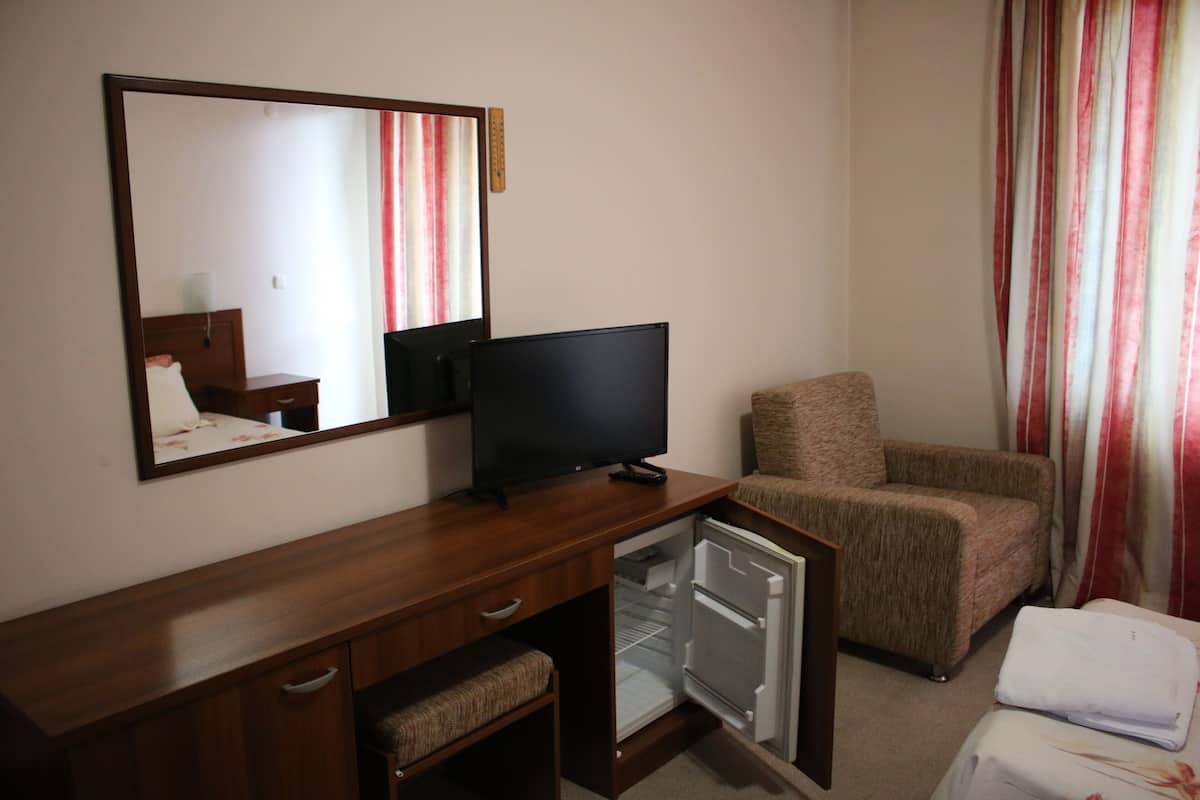 Quarto de hotel com secretária de madeira, televisão, mini-geladeira, poltrona acolchoada e cama com cobertura branca.