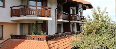 A multi-story building with balconies, terracotta roof tiles, and a tree in the foreground.