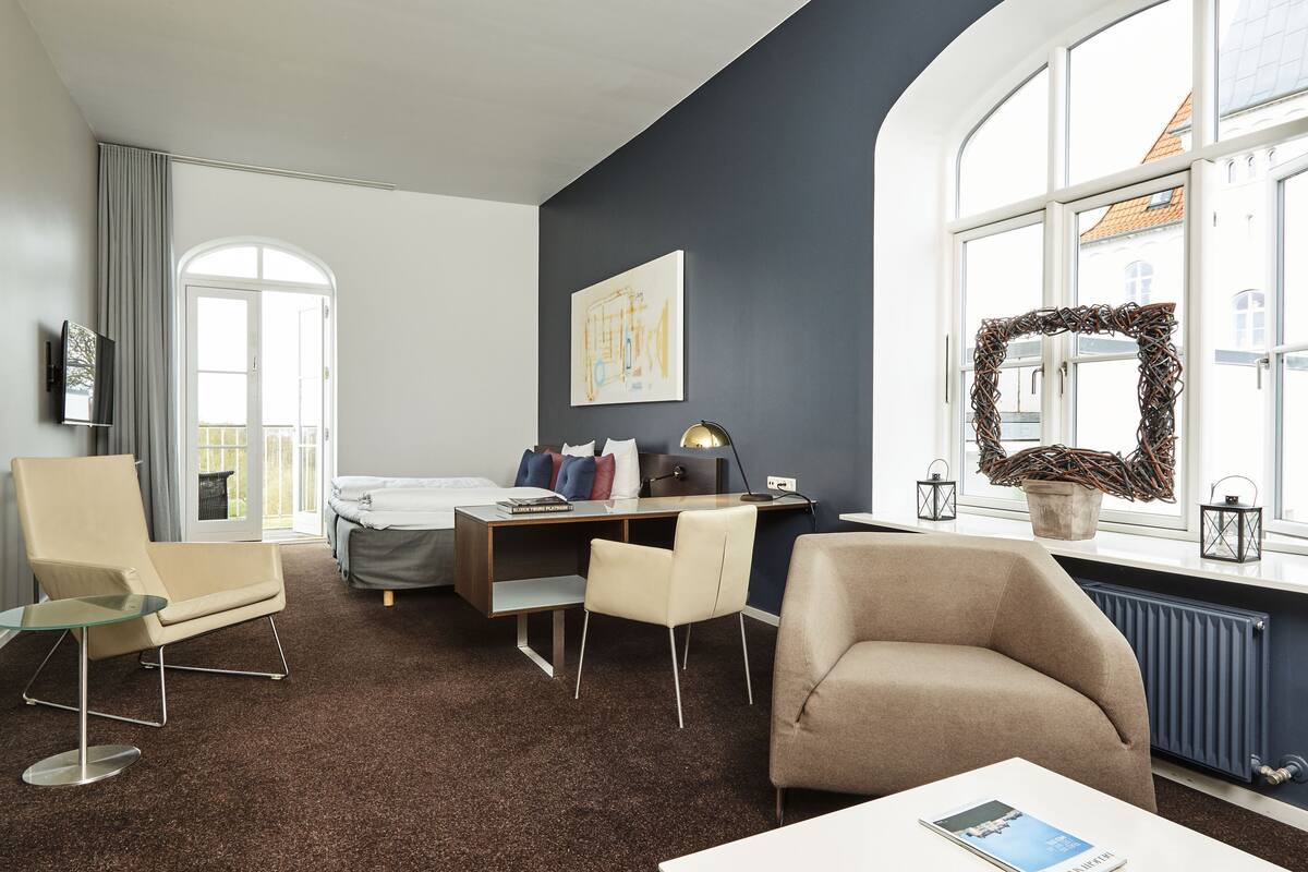 Una habitación de hotel moderna con cama, escritorio, silla y sofá. Una ventana grande ofrece vistas de edificios.