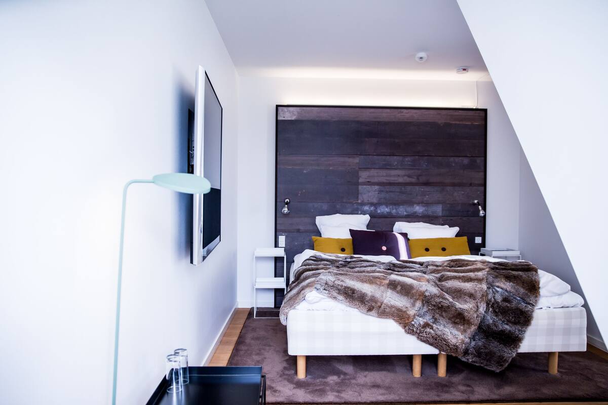 Un dormitorio moderno con techo inclinado, una cama grande con una manta de piel, una mesita de noche, una lámpara de pie y un televisor montado en la pared.