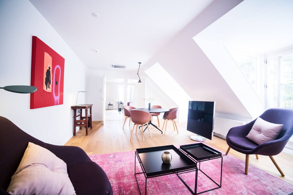 Una sala de estar moderna con un sofá negro, una obra de arte roja, una zona de comedor con una mesa redonda y sillas, y un televisor grande.