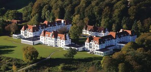 Um edifício grande e com vários andares, com fachada simétrica, rodeado por vegetação exuberante e árvores.
