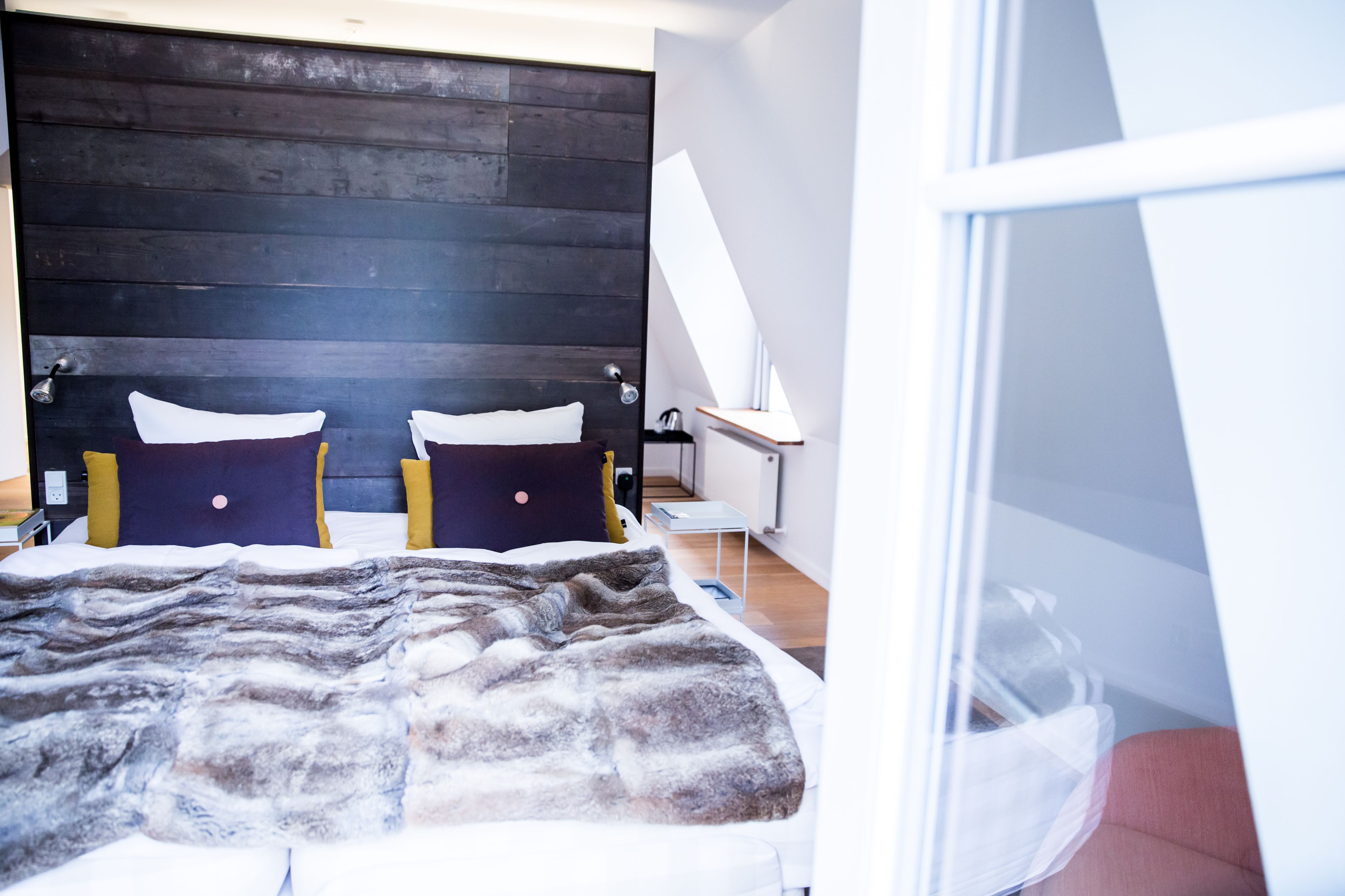 A modern bedroom with a wooden headboard, a bed with a grey and white blanket, and a small bedside table with a lamp.