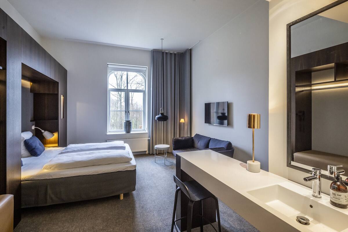 Una habitación de hotel moderna con una cama grande, un escritorio con espejo y una ventana con cortinas.